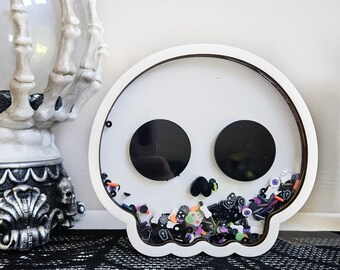 Pode incluir: Um shaker branco em forma de caveira com recortes pretos para os olhos e o nariz. O shaker é preenchido com confetes coloridos com tema de Halloween. Uma mão de esqueleto decorativa e uma bola de cristal estão no fundo.