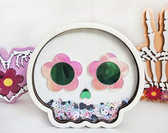 Pode incluir: Uma decoração em forma de caveira de madeira branca com olhos em forma de flor rosa e um nariz em forma de coração verde. A caveira é preenchida com confetes coloridos. Outros itens decorativos estão no fundo.