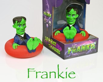 Könnte beinhalten: Grüne Frankenstein-Gummiente in einem roten aufblasbaren Ring. Die Ente trägt einen blauen Anzug und hat ein grünes Gesicht. Die Ente sitzt mit hochgestellten Füßen im Ring. Die Ente ist auf der Schachtel mit "Frankie" beschriftet.