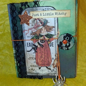 May include: A handmade journal with a green and black cover featuring a vintage-style illustration of a person in a witch hat holding a black cat. The cover includes black lace trim, a glittery orange star, a spider charm, and the text Just A Little Witchy.