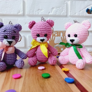 Pode incluir: Três porta-chaves de ursinho de peluche feitos à mão em croché, nas cores roxo, rosa escuro e rosa claro. Cada urso tem olhos de segurança pretos, um nariz preto bordado e uma fita de cetim atada ao pescoço, respetivamente rosa claro, amarelo e verde.