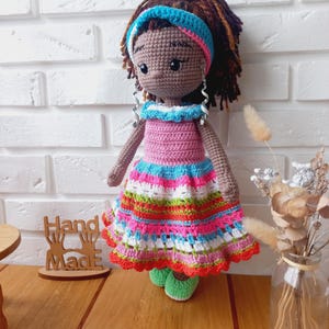 Op de afbeelding: Gehaakte pop met bruine huid, bruin haar en een kleurrijk gestreept jurkje. De pop draagt een blauw hoofdband en zilveren oorbellen. De pop staat op een houten oppervlak met een bord waarop "Hand Made" staat.