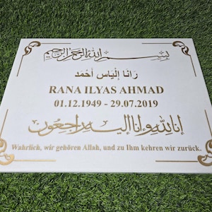 Puede incluir: Placa conmemorativa rectangular blanca con escritura árabe dorada y texto en inglés. El texto incluye el nombre "RANA ILYAS AHMAD" y las fechas "01.12.1949 - 29.07.2019". Bordes dorados decorativos enmarcan el texto.
