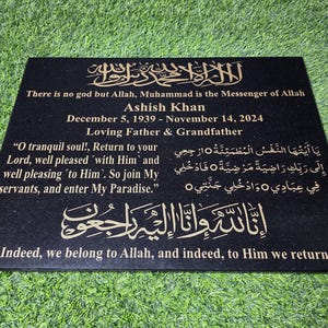 May include: A black granite memorial plaque with gold lettering. The text includes religious phrases, the name Ashish Khan, birth and death dates, and a loving message. The plaque is rectangular.