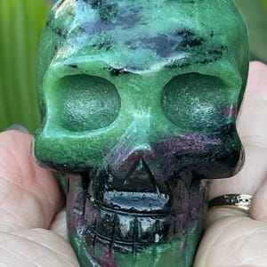 Könnte beinhalten: Eine geschnitzte Totenkopf-Figur aus grünem und lila Stein. Der Schädel hat detaillierte Merkmale, einschließlich Augenhöhlen, einer Nasenhöhle und einem grinsenden Mund. Der Stein hat eine polierte, glänzende Oberfläche, und der Schädel wird in den Händen einer Person gehalten.