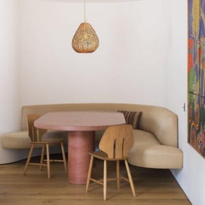 May include: A dining area featuring a round, pink table and two wooden chairs. A beige curved bench is built into the wall. A woven pendant light hangs above the table. A colourful painting hangs on the wall.