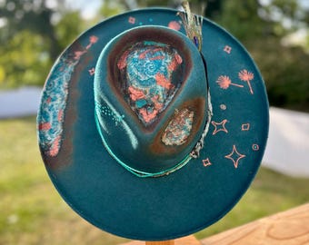 May include: Teal-colored fedora hat with a decorative brim and crown. The hat features a unique painted design in shades of brown, teal, and coral. A peacock feather and beaded trim add detail. The hat is displayed on a wooden stand.