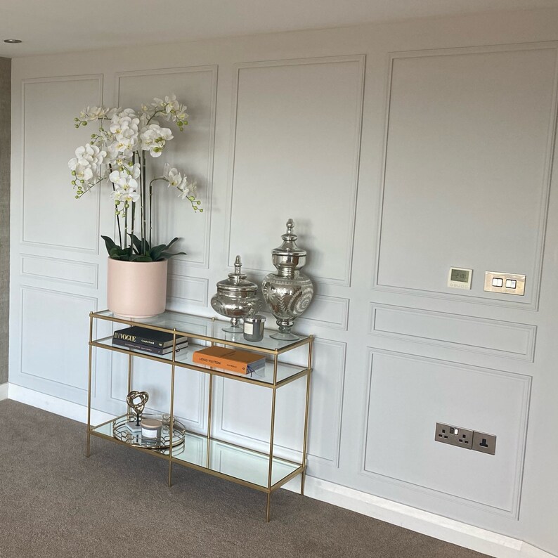 Puede incluir: Una mesa consola de metal dorado con dos estantes de vidrio, decorada con una planta de orqu&iacute;dea blanca en una maceta rosa, dos jarrones decorativos plateados y un libro. La mesa est&aacute; contra una pared blanca con molduras decorativas.