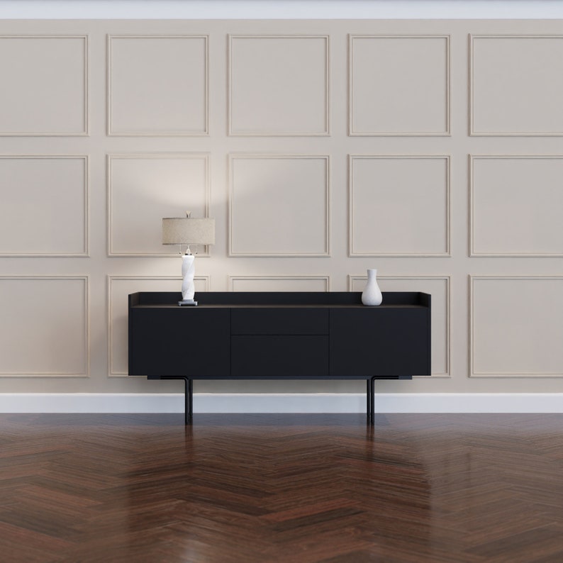 K&ouml;nnte beinhalten: Ein schwarzes Sideboard mit zwei T&uuml;ren und einer Schublade, darauf eine wei&szlig;e Vase und eine Lampe mit einem beigefarbenen Lampenschirm. Das Sideboard steht vor einer Wand mit einem beigefarbenen Wandpaneel-Muster.