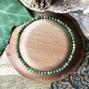 May include: A green beaded bracelet composed of small, round beads. The bracelet is presented on a small, round wooden plate. The background features a piece of driftwood, a green leaf, and a wooden surface.