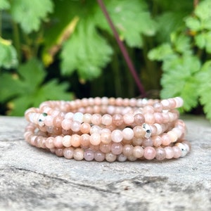May include: A collection of peach-coloured beaded bracelets, each strand composed of small, round beads. The bracelets are interspersed with silver-coloured beads. They are displayed on a grey, textured surface, with green foliage in the background.