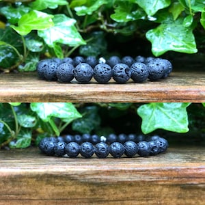 May include: Two black lava stone bead bracelets. The bracelets are displayed on a wooden surface with green foliage in the background. The beads have a porous texture and are strung together with a silver-coloured spacer bead.