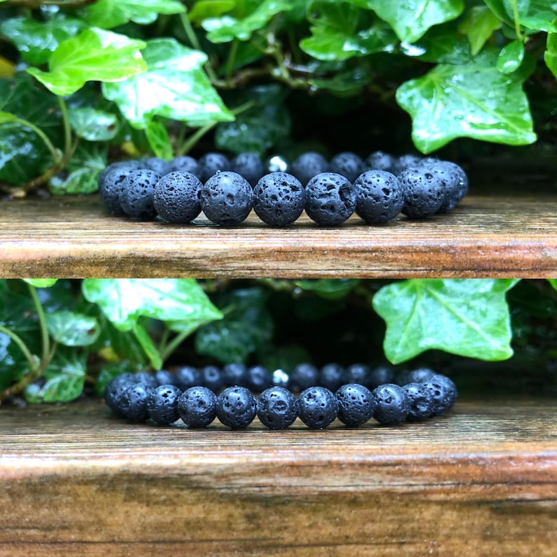Pode incluir: Duas pulseiras de contas de pedra de lava preta. As pulseiras s&atilde;o exibidas em uma superf&iacute;cie de madeira com folhagem verde ao fundo. As contas t&ecirc;m uma textura porosa e s&atilde;o enfiadas com uma conta espa&ccedil;adora prateada.