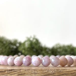 Puede incluir: Un brazalete de cuentas de piedra rosa. Las cuentas son una mezcla de tonos rosas y blancos. El brazalete se exhibe sobre una superficie de madera, con un fondo verde borroso.