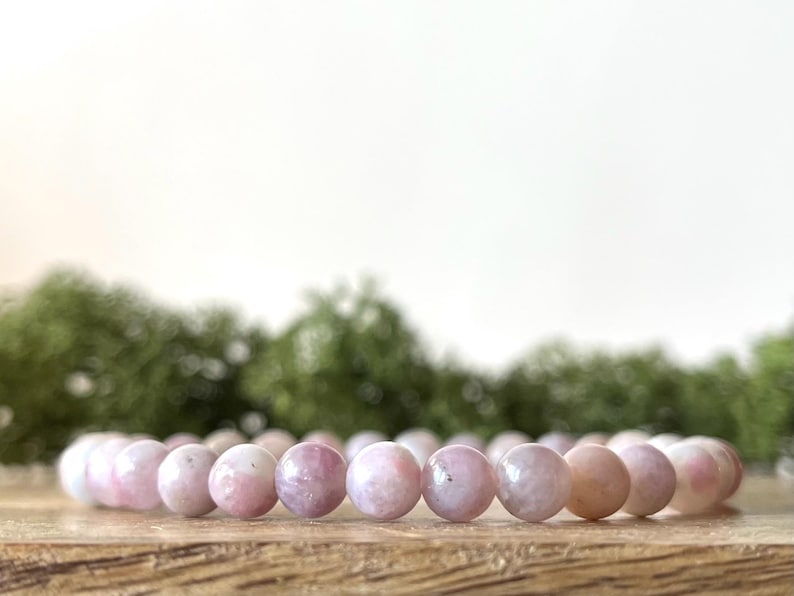 Puede incluir: Un brazalete de cuentas de piedra rosa. Las cuentas son una mezcla de tonos rosas y blancos. El brazalete se exhibe sobre una superficie de madera, con un fondo verde borroso.