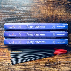 May include: Three boxes of "Fairy Dreams" incense sticks with a purple and white design, and a bundle of incense sticks with red tips. The boxes are stacked on a dark wooden surface.