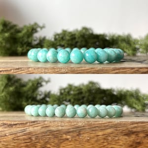May include: A turquoise beaded bracelet, featuring smooth, spherical stones. The bracelet is presented on a wooden surface, with green foliage in the background. The beads are uniform in size and colour, offering a simple, elegant design.