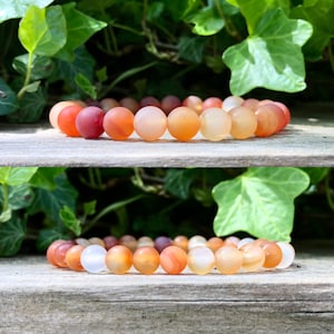 May include: Two bracelets featuring round, polished beads in shades of orange, peach, and white. The bracelets are displayed on a weathered wooden surface, with green foliage in the background. The beads appear to be approximately 1 cm in diameter.