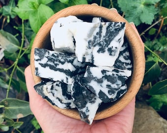 Puede incluir: Un cuenco de madera lleno de piedras de jaspe cebra, con un llamativo patrón en blanco y negro. Dos pulseras con cuentas similares en blanco y negro son visibles debajo del cuenco. El fondo incluye follaje verde.