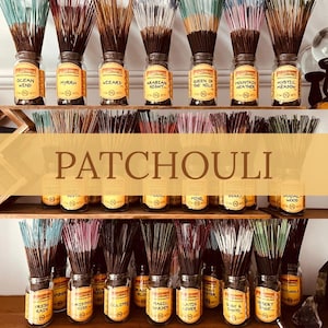 May include: Incense sticks in glass jars with labels, arranged on wooden shelves. Labels include "Ocean Wind," "Myrrh," and "Patchouli." The incense sticks are in various colors. The image includes the word "Patchouli."
