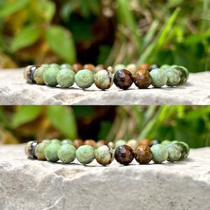 May include: A bracelet made of green, brown, and dark brown faceted beads. The bracelet is presented on a light-coloured stone surface, with a blurred green background.