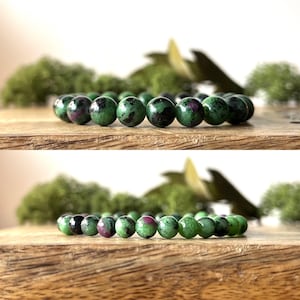 May include: A bracelet made of green and dark red gemstone beads. The round beads are a mix of green with dark red inclusions. The bracelet is displayed on a wooden surface with a blurred green background.