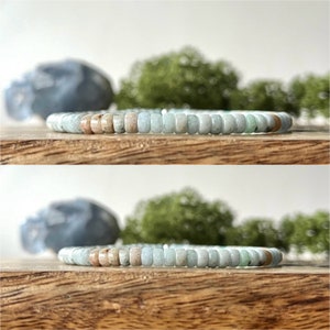 May include: A beaded bracelet featuring small, rectangular beads in shades of light blue, tan, and green. The bracelet is displayed on a wooden surface, with a blurred background of greenery and a blue stone.