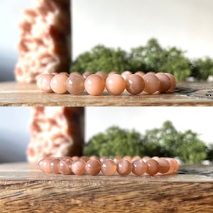 May include: A peach-coloured beaded bracelet. The bracelet is made of round, polished beads. The beads have a smooth surface and a warm, peachy hue. The bracelet is displayed on a wooden surface.