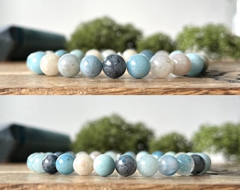 Puede incluir: Una pulsera de cuentas esféricas en tonos azul claro, crema y gris oscuro. La pulsera se muestra sobre una superficie de madera, con un fondo verde borroso.