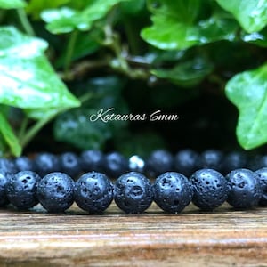 Pode incluir: Uma pulseira de pedra de lava preta com contas de 6 mm. A pulseira &eacute; feita com um cord&atilde;o el&aacute;stico preto. O texto "Katauras 6mm" &eacute; vis&iacute;vel na imagem. O fundo apresenta folhagem verde.