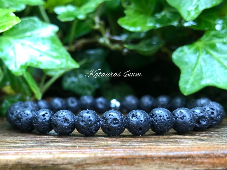 Pode incluir: Uma pulseira de pedra de lava preta com contas de 6 mm. A pulseira &eacute; feita com um cord&atilde;o el&aacute;stico preto. O texto "Katauras 6mm" &eacute; vis&iacute;vel na imagem. O fundo apresenta folhagem verde.