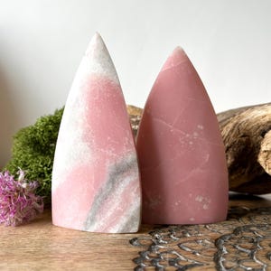 May include: Two pink gemstone obelisks. One is a solid, soft pink, while the other has white and gray veining. They are displayed on a wooden surface with moss and flowers, creating a natural aesthetic.