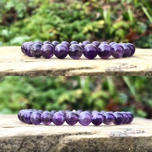 May include: A bracelet featuring polished, round amethyst beads. The beads are a deep purple hue and strung together. The bracelet is displayed on a stone surface with a blurred green backdrop.