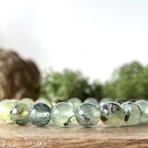 May include: A Prehnite gemstone bracelet featuring translucent, pale green beads with dark inclusions. The round beads are strung together, resting on a wooden surface. The background includes blurred greenery and a light-colored backdrop.