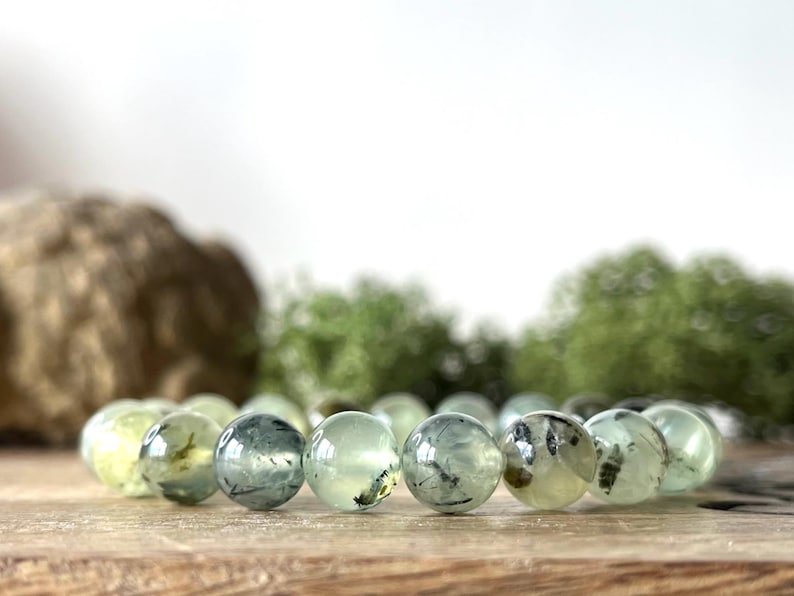 May include: A Prehnite gemstone bracelet featuring translucent, pale green beads with dark inclusions. The round beads are strung together, resting on a wooden surface. The background includes blurred greenery and a light-colored backdrop.