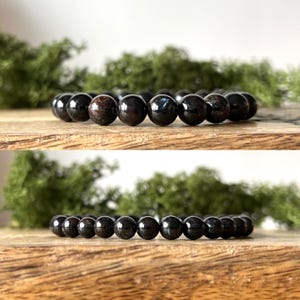 May include: A black beaded bracelet with a polished finish. The bracelet is made of round, dark beads, likely gemstones, strung together on an elastic band. The beads have a glossy sheen, reflecting light. The bracelet is displayed on a wooden surface.