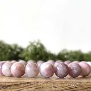 Puede incluir: Un brazalete de cuentas redondas y pulidas, en tonos rosa y blanco. El brazalete se exhibe sobre una superficie de madera, con un fondo verde borroso.