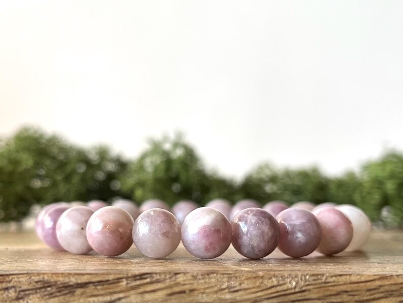 Puede incluir: Un brazalete de cuentas redondas y pulidas, en tonos rosa y blanco. El brazalete se exhibe sobre una superficie de madera, con un fondo verde borroso.