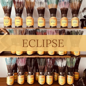 May include: A display of incense sticks in glass jars, labeled with names like "Ocean Wind" and "Myrrh." The jars are arranged on wooden shelves, with the word "ECLIPSE" prominently displayed. The incense sticks have various colors.
