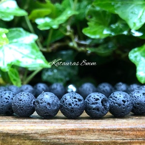 Pode incluir: Uma pulseira de pedra de lava preta com contas de 8 mm. A textura porosa das contas &eacute; vis&iacute;vel. A pulseira &eacute; exibida em uma superf&iacute;cie de madeira, com folhagem verde ao fundo. O texto "Katauras 8mm" &eacute; vis&iacute;vel.