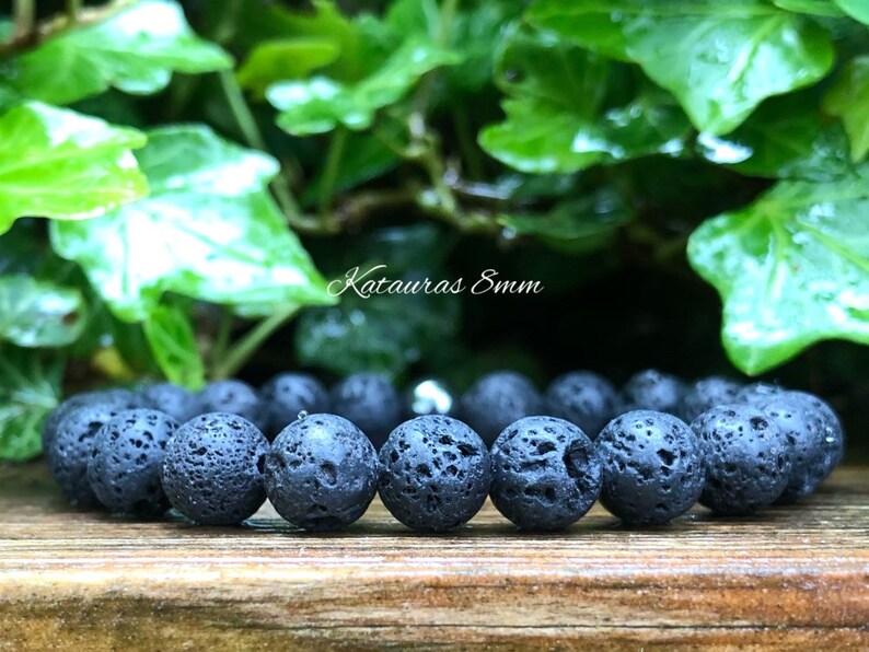 Pode incluir: Uma pulseira de pedra de lava preta com contas de 8 mm. A textura porosa das contas &eacute; vis&iacute;vel. A pulseira &eacute; exibida em uma superf&iacute;cie de madeira, com folhagem verde ao fundo. O texto "Katauras 8mm" &eacute; vis&iacute;vel.