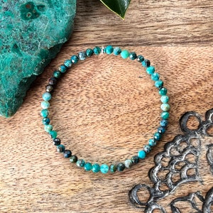 May include: A turquoise beaded bracelet with small, faceted beads. The bracelet is elasticated and sits on a wooden surface. A piece of turquoise stone and a green leaf are in the background.