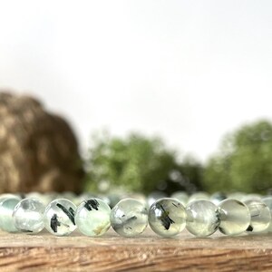 May include: A bracelet featuring translucent, light green beads with dark inclusions. The beads are strung together, resting on a wooden surface. The background includes blurred greenery and a light-colored backdrop.