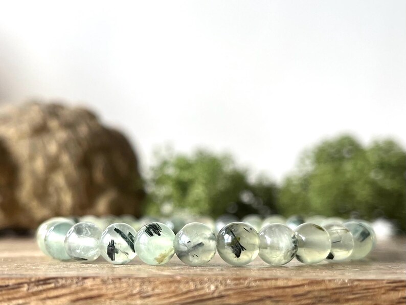 May include: A bracelet featuring translucent, light green beads with dark inclusions. The beads are strung together, resting on a wooden surface. The background includes blurred greenery and a light-colored backdrop.