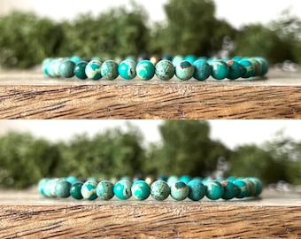 May include: A beaded bracelet featuring turquoise and green jasper stones. The round beads are strung together, showcasing a mix of colors and patterns. The bracelet is displayed on a wooden surface with a blurred green background.