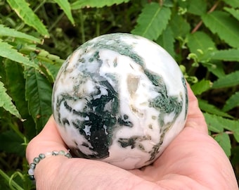 Puede incluir: Una esfera pulida de ágata musgosa con patrones blancos y verdes. La gema se sostiene en una mano, con follaje verde en el fondo. Un objeto decorativo natural.