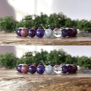 May include: A bracelet made of various gemstone beads in shades of purple, clear, and brown. The bracelet is displayed on a wooden surface with green moss in the background. The beads are round and of varying sizes.