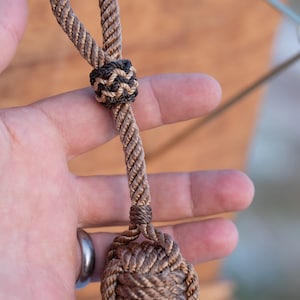 May include: A decorative rope knot, featuring a loop at the top and a large, spherical knot at the bottom. The rope is a mix of tan and brown colors, with a smaller, contrasting knot in black and tan. The item is held in a hand.