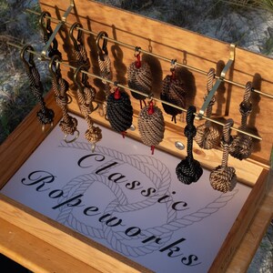 May include: A wooden box with a brass rack holding a variety of rope knots. The box is open and the inside is lined with white paper that says "Classic Ropeworks".