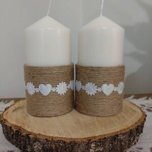 May include: Two white pillar candles with burlap and white lace trim, sitting on a wooden slice.